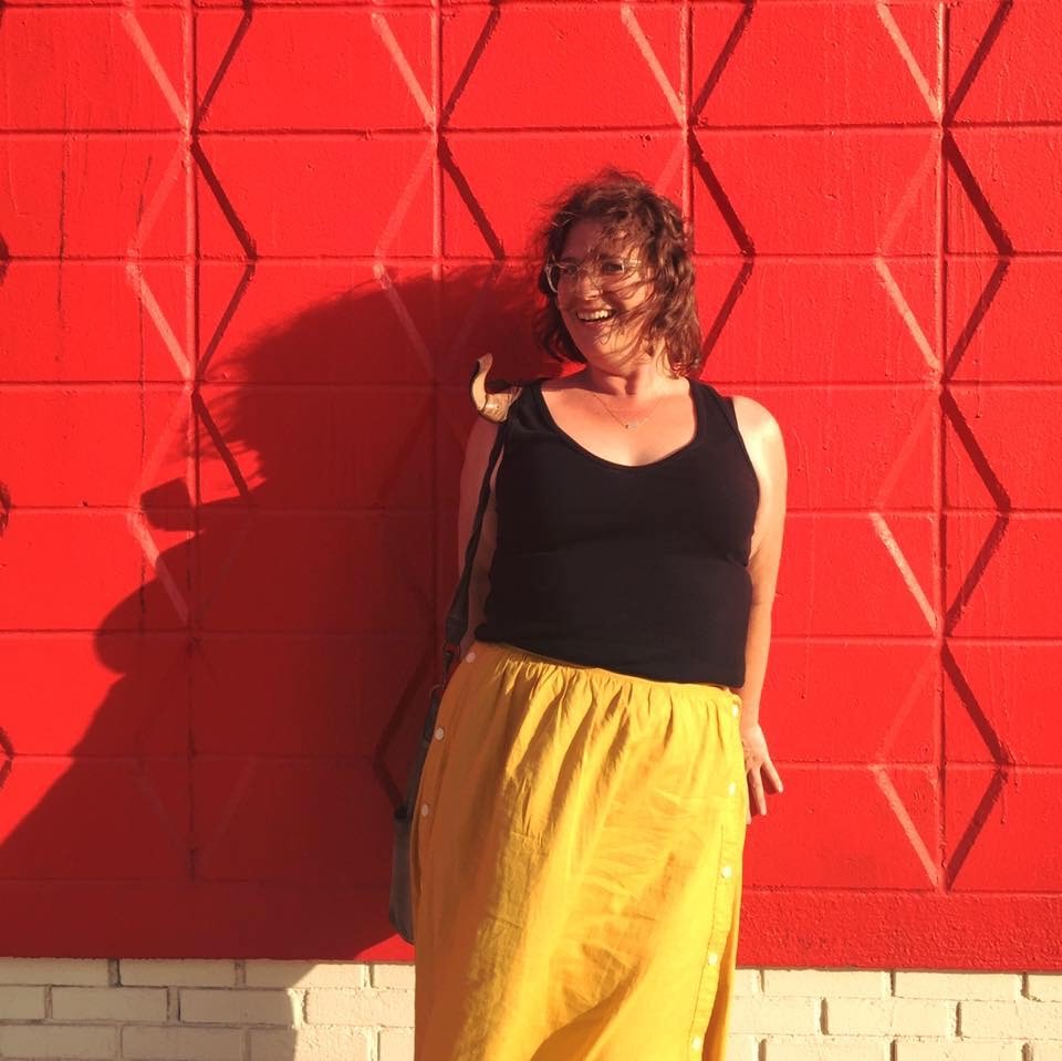 a photo of Nicole Treska standing in front of a red wall wearing a black shirt and yellow dress